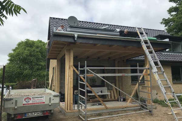 Handwerker führen Dacharbeiten an einem Holzvordach durch, Gerüst und Bauwagen sind im Vordergrund sichtbar.