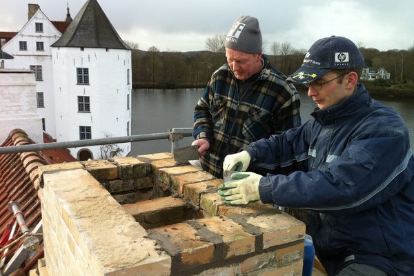 Zwei Handwerker führen Maurerarbeiten an einem Schornstein auf einem Dach durch, im Hintergrund der Schlossteich in Glücksburg.