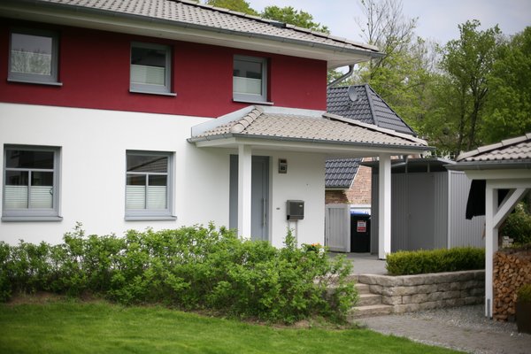 Modernes Einfamilienhaus mit roter Fassade, begrüntem Vorgarten und überdachtem Eingangsbereich.