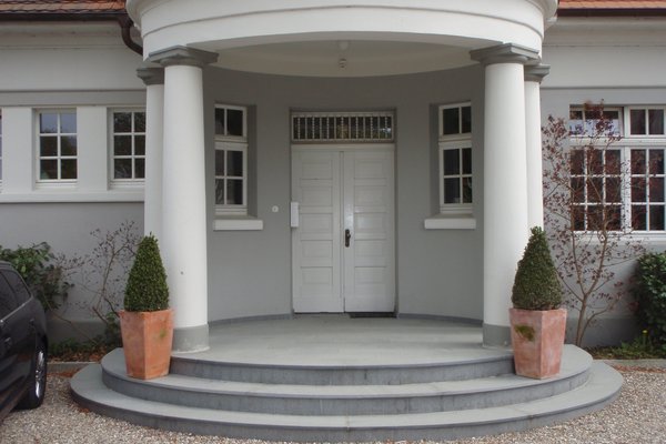 Eingangsbereich mit runder Treppe, Säulen und zwei Topfpflanzen vor einer grauen Fassade.