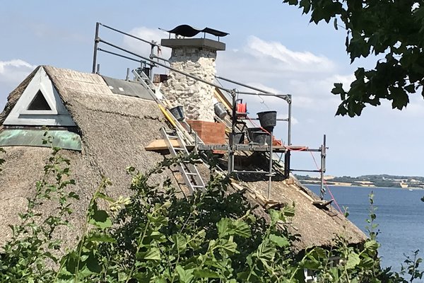 Sanierungsarbeiten an einem Reetdach mit Gerüst und Kamin, Blick auf das Wasser im Hintergrund.