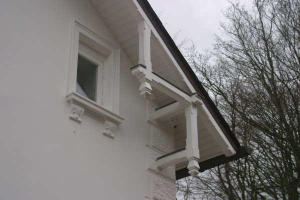 Detailansicht eines weißen Hauses mit dekorativen Fassadenelementen und einem Fenster, umgeben von Bäumen.