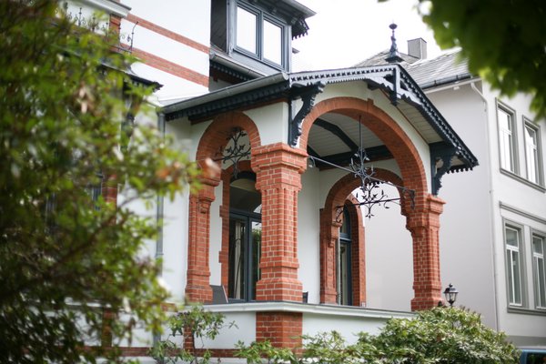 Historische Fassade mit verzierten Säulen und einem Balkon, umgeben von grünen Pflanzen.