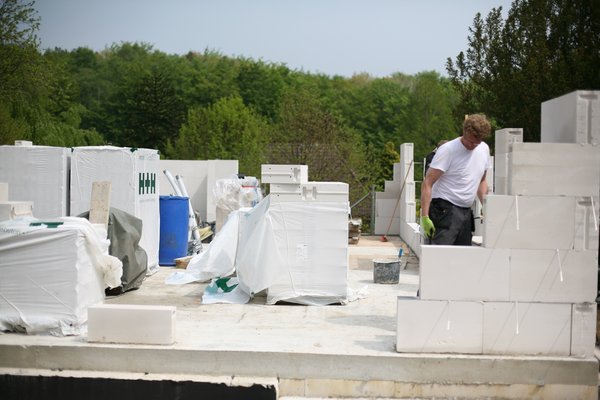 Ein Bauarbeiter arbeitet auf einer Baustelle mit weißen Mauersteinen, umgeben von Baustellenmaterialien und Bäumen.