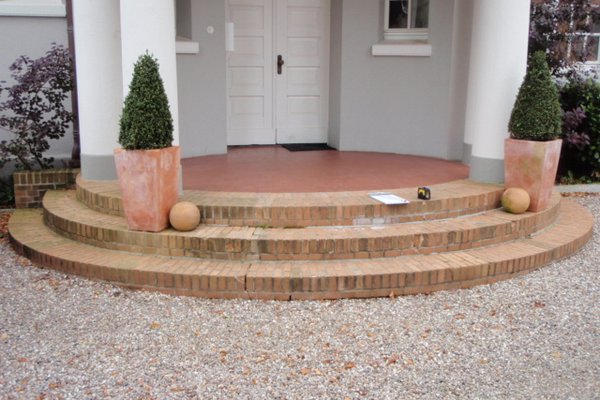 Eingangsbereich mit dreistufiger Treppe aus Ziegelsteinen, bepflanzt mit zwei Kübelpflanzen und dekorativen Kugeln.