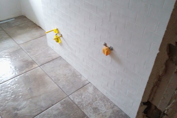 Fliesenarbeiten in einem Badezimmer mit weißen Wandfliesen und grauen Bodenfliesen, sichtbare Wasseranschlüsse.