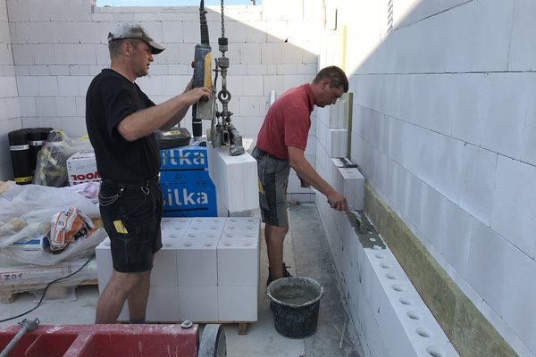 Zwei Handwerker führen Maurerarbeiten mit weißen Blocksteinen durch, einer verputzt, der andere hebt Steine mit einem Kran.