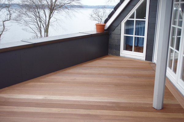 Terrasse mit Holzbelag und Blick auf einen See, umgeben von Bäumen und einem modernen Gebäude.