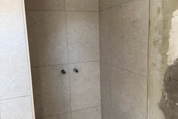 Moderne Duschkabine mit hellen Fliesen und zwei Wasseranschlüssen, unvollendete Wandbearbeitung sichtbar.