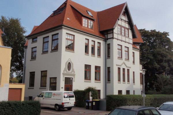 Historisches Gebäude mit rotem Satteldach, weißen Wänden und großen Fenstern, umgeben von Bäumen und Fahrzeugen.
