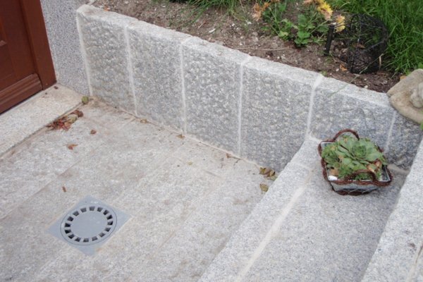 Treppenstufen aus grauem Stein mit einem Gully und einem Pflanzkorb in einer begrünten Umgebung.