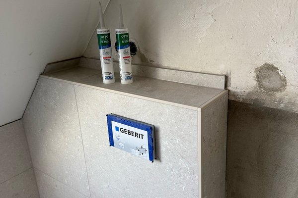 Sanierter Bereich mit Fliesen, einem Geberit-Schild und zwei Tuben Silikon auf einer Ablage.