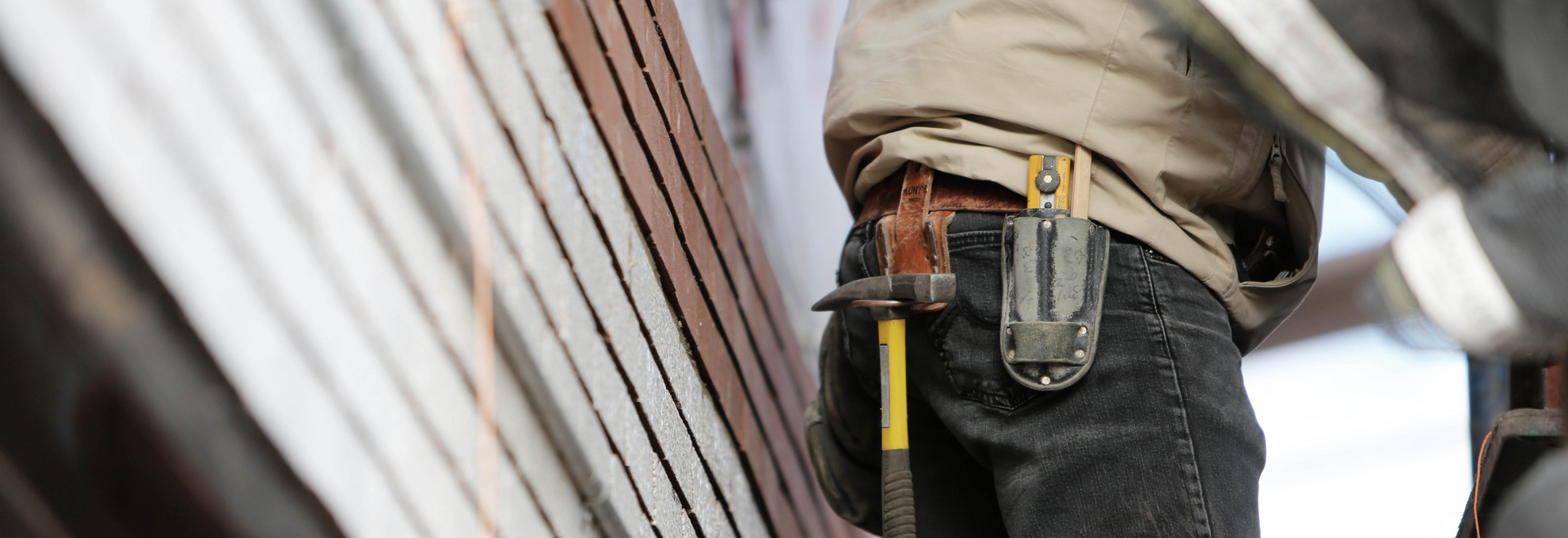 Ein Handwerker steht an einer Baustelle, mit Werkzeugen in der Gürteltasche und Ziegelsteinen im Hintergrund.