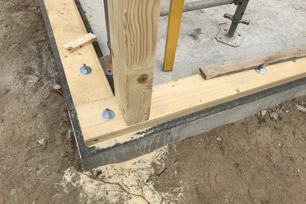 Holzrahmenkonstruktion mit Schrauben an einer Betonfundamentkante, umgeben von unbefestigtem Erdreich.